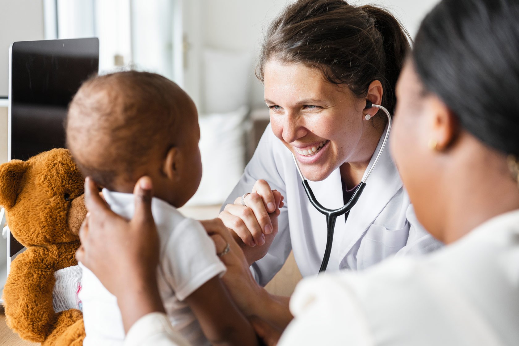 Female doctor holding hand of young boy, with mother and teddy bear in background